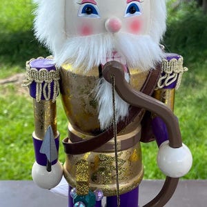 May include: A nutcracker figure wearing a purple hat with a gold crown, a gold and purple uniform with gold trim, and white faux fur hair and beard. The figure holds a brown bow and a spear, standing beside a green tree and a small owl figure.
