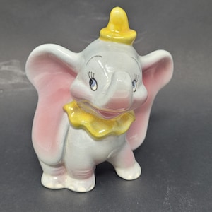 Könnte beinhalten: Eine Keramikfigur von Dumbo dem Elefanten aus Disneys Zeichentrickfilm. Die Figur ist in Grautönen, Rosa und Gelb bemalt. Dumbo hat große rosa Ohren, einen gelben Hut und einen gelben Kragen.