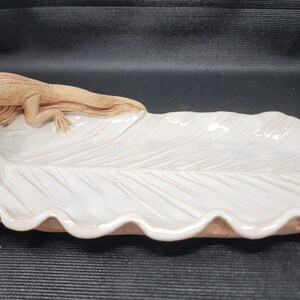 May include: A decorative ceramic leaf-shaped dish with a detailed alligator figurine. The dish is white with a scalloped edge and the alligator is a light brown color. This unique piece is suitable for home decor.