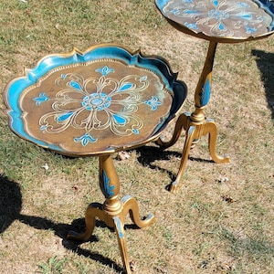 May include: Two ornate side tables with scalloped edges. The tables have a gold finish with teal and gold floral designs on the tabletop and legs. The tables are set on a grassy surface.