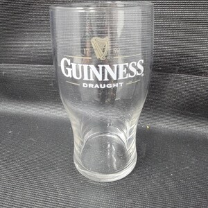 May include: A clear Guinness draught glass with the Guinness logo in white and gold. The glass has a curved shape and a wide top, tapering to a smaller base. The glass is empty.