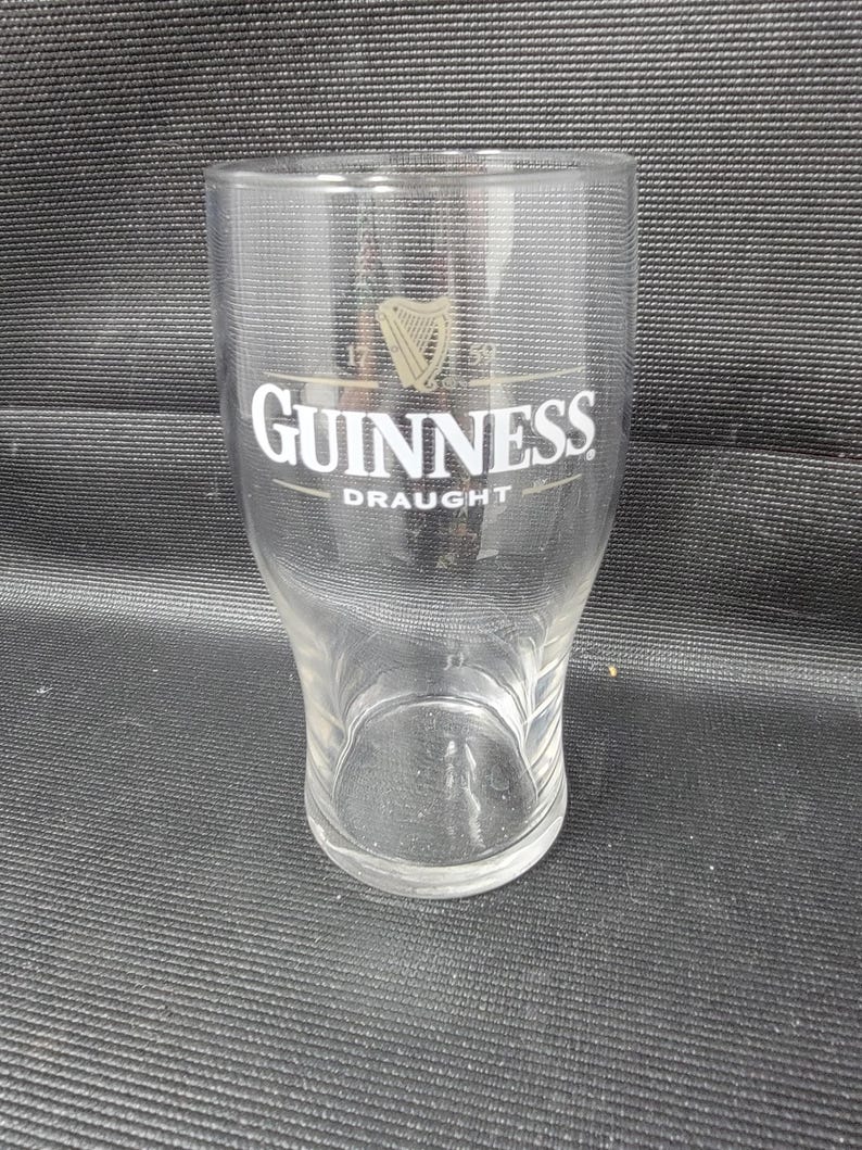 May include: A clear Guinness draught glass with the Guinness logo in white and gold. The glass has a curved shape and a wide top, tapering to a smaller base. The glass is empty.