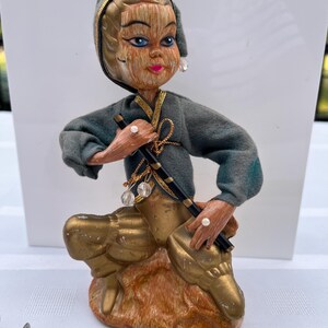 May include: A decorative figurine of a person in a crouching pose, wearing a dark green hooded jacket with gold trim and gold pants. The figure has textured hair, blue eyes, and pink lips, holding a black rod. A yellow measuring tape shows 5.5 inches.