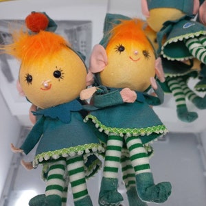 May include: Three plush elf ornaments with orange hair, green and white striped legs, and green felt hats. The elves are wearing green and white striped stockings and have a green felt dress with white trim.