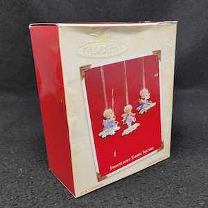 May include: A red box with a white label that says "Keepsake Ornament" and "Frostlight Faerie Sisters". The box contains a set of three small, white, fairy ornaments with blue and purple accents.