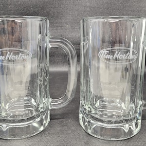 May include: Two clear glass beer mugs with handles. Each mug features the Tim Hortons logo etched on the front. The mugs are rectangular with a thick base and a curved handle. The background is a dark grey surface.