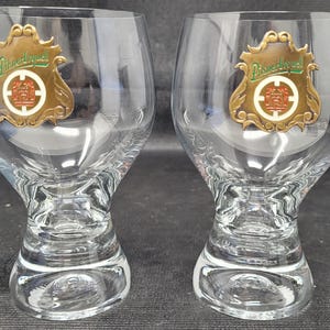 May include: Two clear, stemmed beer glasses with a decorative emblem featuring the text "Pilsner Urquell" in green and gold. The glasses have a unique, textured base and a wide bowl.