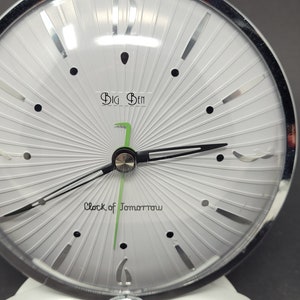 May include: A white clock with a silver rim and black hands. The clock face has a sunburst design and the text "Big Ben Clock of Tomorrow" in black lettering.