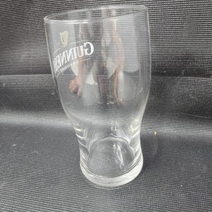 May include: A clear, curved pint glass with the Guinness logo in white. The glass has a wide top, a narrow waist, and a flared base. The Guinness logo includes a harp symbol and the word "Guinness".
