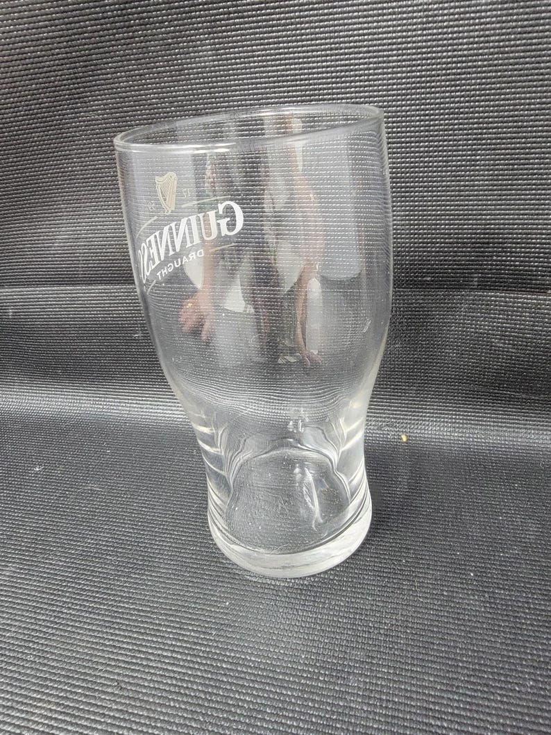 May include: A clear, curved pint glass with the Guinness logo in white. The glass has a wide top, a narrow waist, and a flared base. The Guinness logo includes a harp symbol and the word "Guinness".