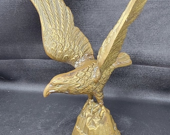 May include: A bronze-colored eagle sculpture with outstretched wings, perched atop a textured rock base. The eagle's feathers are detailed, and the sculpture has a metallic sheen. The wingspan is wide, and the eagle appears to be in flight.