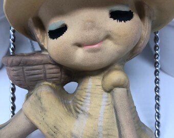 May include: A ceramic figurine of a girl wearing a straw hat and a striped dress. She is holding a basket in her hand and has a sweet expression on her face.