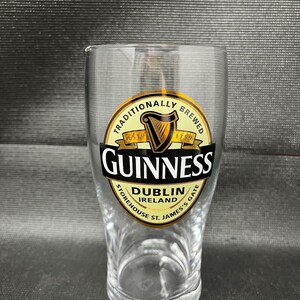May include: A clear pint glass with the Guinness logo. The logo is a gold and black circular design with the word "Guinness" in bold white letters. The glass also features the text "Dublin Ireland" and "St. James's Gate".