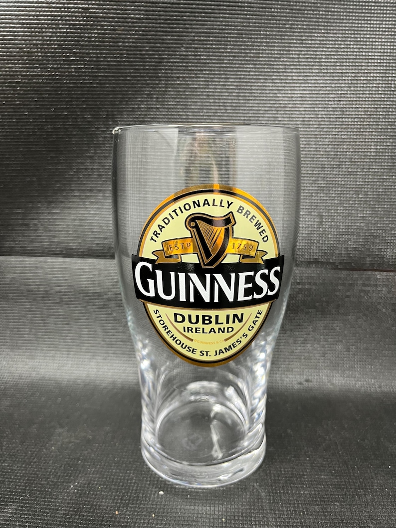 May include: A clear pint glass with the Guinness logo. The logo is a gold and black circular design with the word "Guinness" in bold white letters. The glass also features the text "Dublin Ireland" and "St. James's Gate".