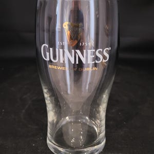May include: A clear Guinness pint glass with the Guinness logo in white and gold. The glass is 6 inches tall and 3.25 inches in diameter. The text "BREWED IN DUBLIN" is printed below the logo.