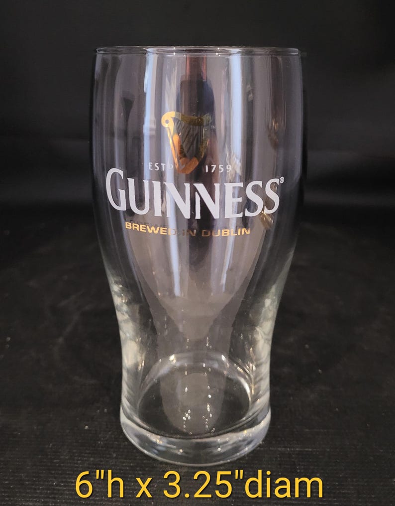 May include: A clear Guinness pint glass with the Guinness logo in white and gold. The glass is 6 inches tall and 3.25 inches in diameter. The text "BREWED IN DUBLIN" is printed below the logo.