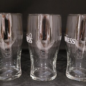 May include: Three clear pint glasses with the word "Guinness" printed in white. The glasses are empty and have a curved shape, with a wider top and a narrower base. The background is dark.