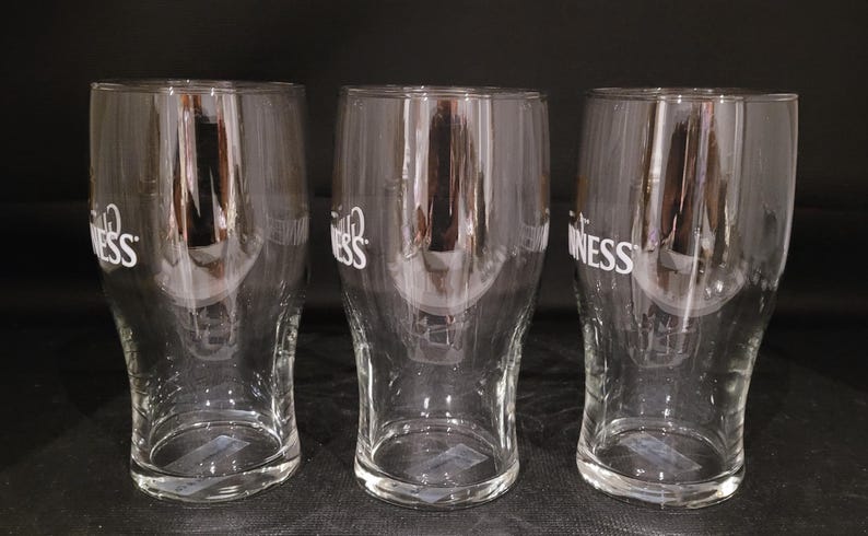 May include: Three clear pint glasses with the word "Guinness" printed in white. The glasses are empty and have a curved shape, with a wider top and a narrower base. The background is dark.