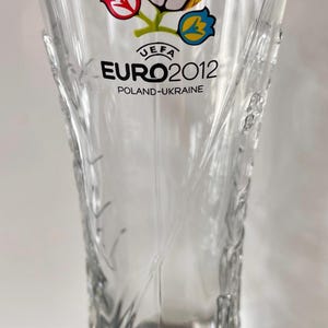 May include: Clear glass pint glass with the UEFA Euro 2012 logo. The logo features a soccer ball and the text "EURO 2012 POLAND-UKRAINE". The glass has a decorative etched design on the sides.