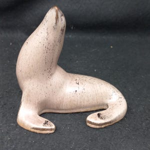 May include: A ceramic seal figurine in a light brown colour with a speckled finish. The seal is sitting on its belly with its head raised.