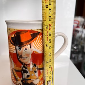 K&ouml;nnte beinhalten: Eine wei&szlig;e Keramik-Tasse mit einem Bild der Figur Woody aus dem Film Toy Story. Die Tasse ist etwa 10 cm hoch und hat einen gebogenen Henkel. Das Design ist farbenfroh.