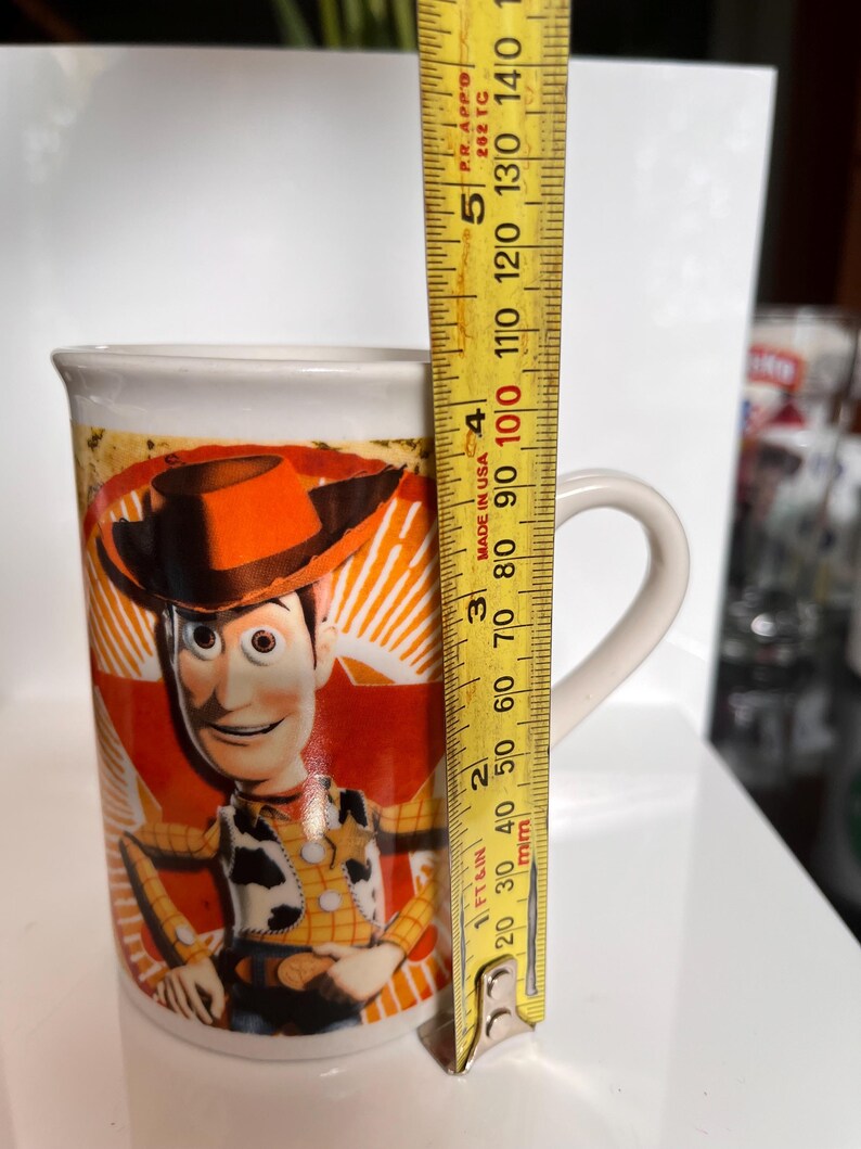 K&ouml;nnte beinhalten: Eine wei&szlig;e Keramik-Tasse mit einem Bild der Figur Woody aus dem Film Toy Story. Die Tasse ist etwa 10 cm hoch und hat einen gebogenen Henkel. Das Design ist farbenfroh.