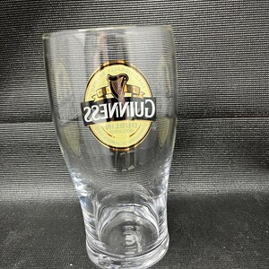 May include: A clear, tulip-shaped pint glass with the Guinness logo. The logo is a gold emblem with a harp and the word "Guinness" in a black, bold font. The glass is empty and sits on a dark surface.