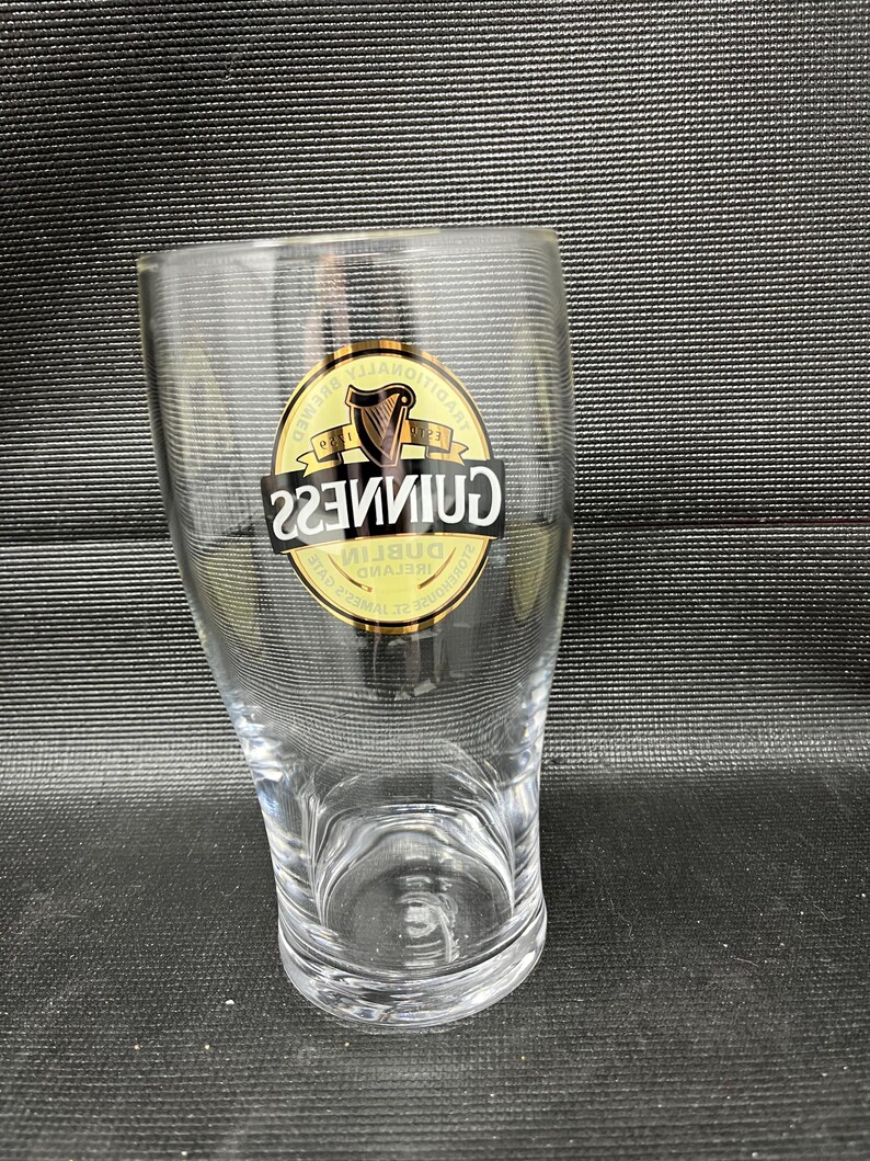 May include: A clear, tulip-shaped pint glass with the Guinness logo. The logo is a gold emblem with a harp and the word "Guinness" in a black, bold font. The glass is empty and sits on a dark surface.