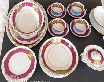 Könnte beinhalten: Ein Vintage-Porzellan-Dinner-Set mit einem burgunderfarbenen und goldenen Blumendesign. Das Set beinhaltet eine große ovale Servierplatte, eine kleinere runde Servierschüssel, vier Speiseteller, vier Teetassen und Untertassen sowie eine abgedeckte Servierschüssel.