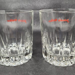 May include: Two clear, faceted drinking glasses with a red logo. The glasses have a thick base and a textured, vertical pattern on the sides. The glasses are made of clear glass and are set against a dark background.