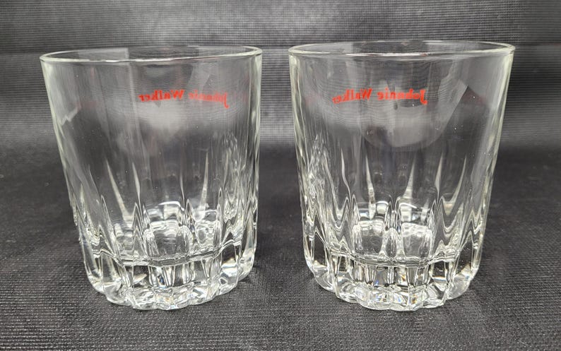 May include: Two clear, faceted drinking glasses with a red logo. The glasses have a thick base and a textured, vertical pattern on the sides. The glasses are made of clear glass and are set against a dark background.