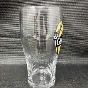 May include: A clear, cylindrical pint glass with a slightly flared top. A gold and black label with the word "Guinness" is affixed to the side. The glass is empty and sits on a dark surface.