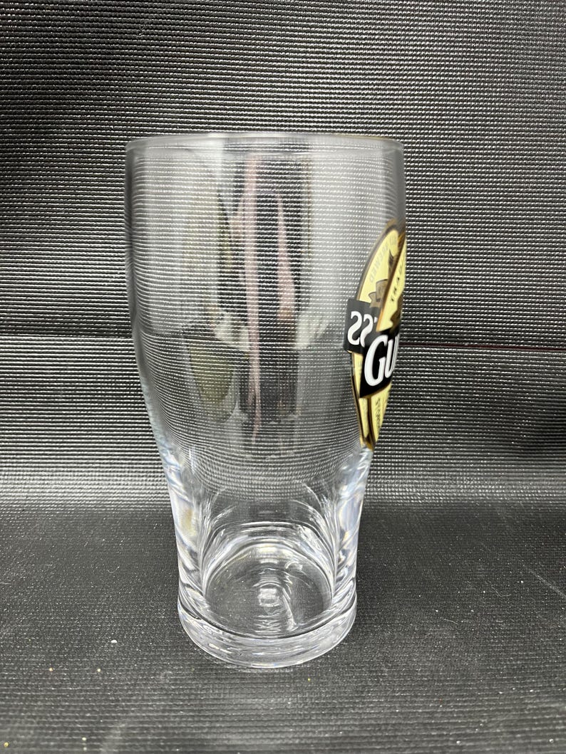 May include: A clear, cylindrical pint glass with a slightly flared top. A gold and black label with the word "Guinness" is affixed to the side. The glass is empty and sits on a dark surface.