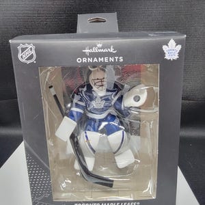 Peut inclure: Ornement de Noël des Maple Leafs de Toronto dans une boîte. L'ornement est une figurine de gardien de but de hockey en uniforme bleu et blanc. La boîte porte les logos de la LNH et de Hallmark Ornaments.