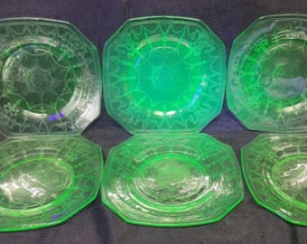 May include: Six green glass plates with a decorative etched design. The plates are square with rounded corners.
