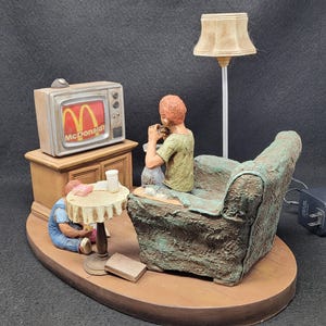 Op de afbeelding: Een keramiek beeldje dat een gezin laat zien dat televisie kijkt. De vader zit op een bank met een lamp achter hem. De moeder zit op de vloer met een kind. De televisie toont een commercial voor McDonald's. Het beeldje is geschilderd in realistische kleuren en details.