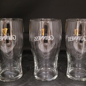 May include: Three clear pint glasses with the Guinness logo in white. The logo includes a harp symbol. The glasses are empty and have a curved shape, wider at the top and bottom. The glasses are on a dark surface.