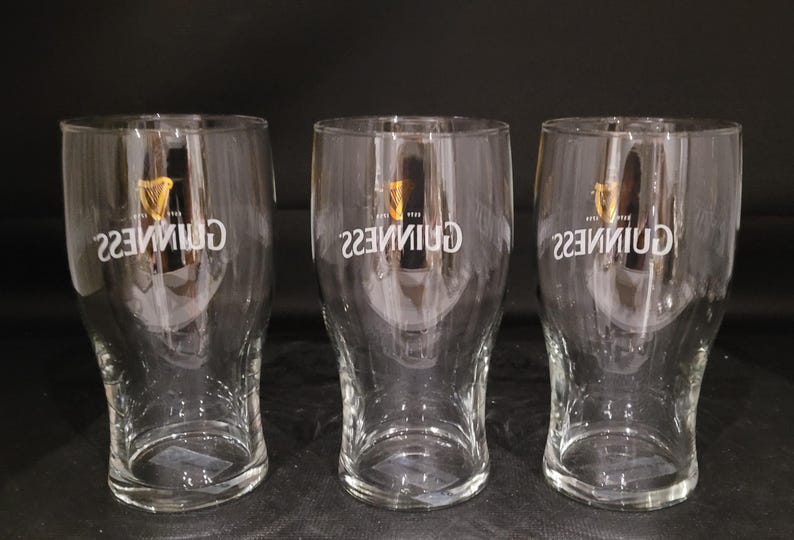 May include: Three clear pint glasses with the Guinness logo in white. The logo includes a harp symbol. The glasses are empty and have a curved shape, wider at the top and bottom. The glasses are on a dark surface.