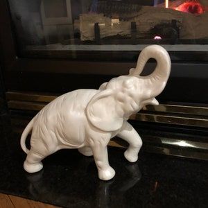 Könnte beinhalten: Weiße Keramik-Elefantenfigur mit erhobenem Rüssel. Der Elefant steht auf einer schwarzen Oberfläche.