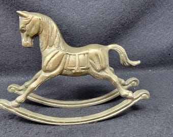 Puede incluir: Una figurita de caballo de balancín de latón con un diseño detallado. El caballo está en una pose de galope con la cabeza girada hacia un lado. La base de balancín está hecha de dos piezas curvas de latón.