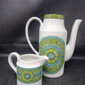 Pode incluir: Conjunto de bule e cremeira em cerâmica branca. O bule tem uma tampa verde e um grande desenho circular em tons de verde e azul. O creme tem um design correspondente. Em um fundo escuro.