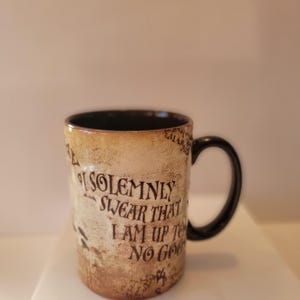 May include: A ceramic mug with a black handle and interior. The exterior has a parchment-like appearance with the phrase "I SOLEMNLY SWEAR THAT I AM UP TO NO GOOD" in black lettering. The mug is a standard size.