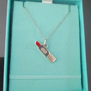 May include: A silver necklace with a lipstick charm, presented in a light blue box. The lipstick charm has a red top and a silver body with vertical lines. The necklace is displayed on a light blue velvet-like material.