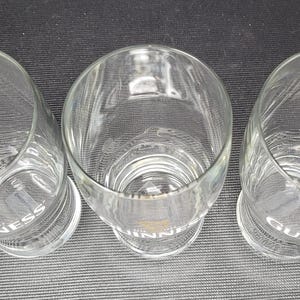 May include: Three clear, cylindrical drinking glasses with a slightly flared top. The word "GUINNESS" is printed in white on the base of each glass. The glasses are empty and appear to be made of glass.