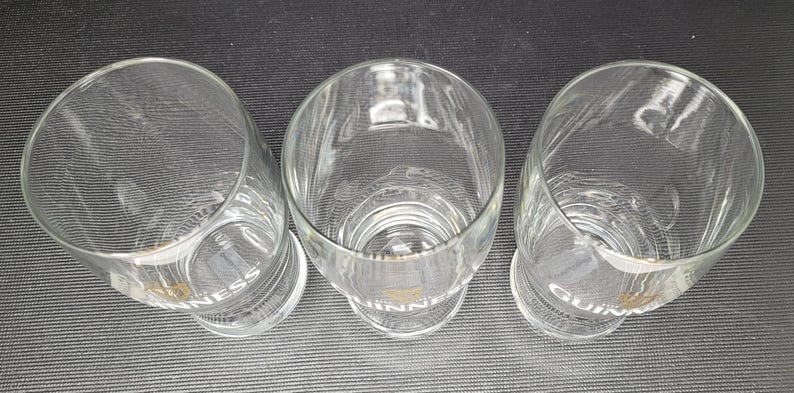 May include: Three clear, cylindrical drinking glasses with a slightly flared top. The word "GUINNESS" is printed in white on the base of each glass. The glasses are empty and appear to be made of glass.