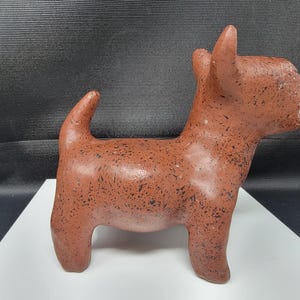 Pode incluir: Estatueta de cão em terracota com manchas pretas. A escultura tem uma forma estilizada com orelhas direitas e cauda enrolada. O cão está em pé sobre uma superfície branca contra um fundo escuro.