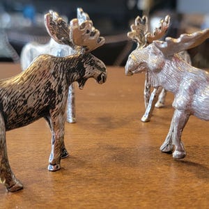 May include: Three silver moose figurines with detailed antlers are displayed on a wooden surface. The moose are in various poses, with one facing the viewer and another facing the first. The figurines have a metallic finish.