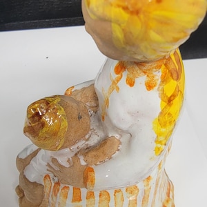May include: A ceramic figurine of a seated figure with yellow hair and a white dress. The dress is decorated with orange stripes and accents. The figure is holding a brown object. The figurine is handmade and has a unique, artistic style.