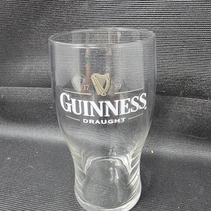 May include: A clear Guinness draught glass with the Guinness logo in white and gold. The glass has a wide base and tapers inward towards the top. The text "Guinness Draught" is printed on the glass.