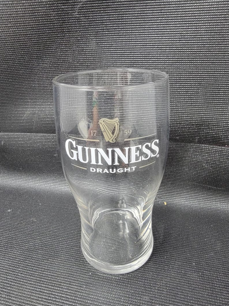 May include: A clear Guinness draught glass with the Guinness logo in white and gold. The glass has a wide base and tapers inward towards the top. The text "Guinness Draught" is printed on the glass.